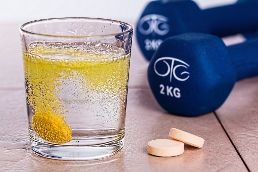 Prozirna čaša s gaziranim napitkom i šumećom tabletom, pored koje se nalaze dvije bijele tablete i plave bučice od 2 kg.